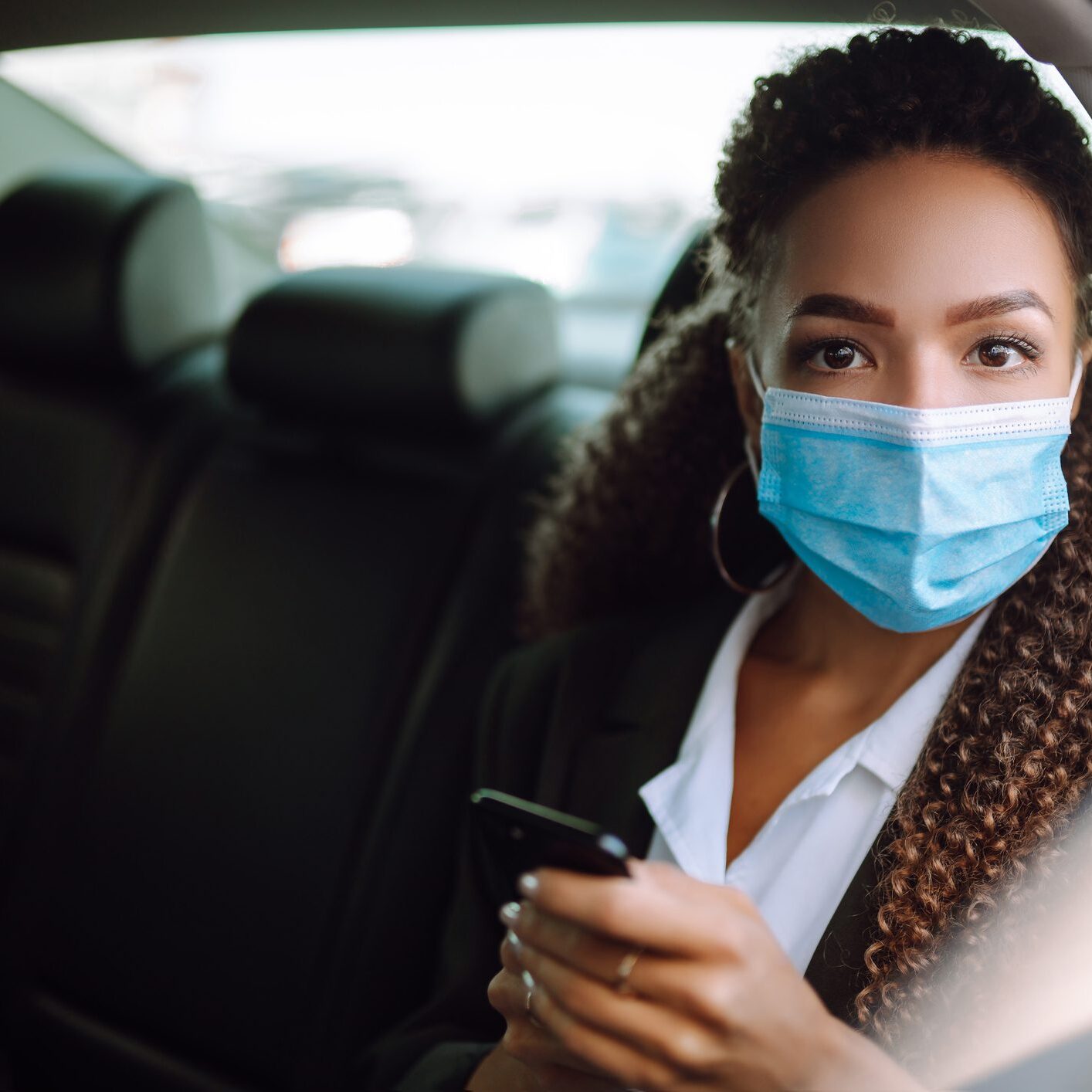 A woman in a car wearing a mask and holding her phone.